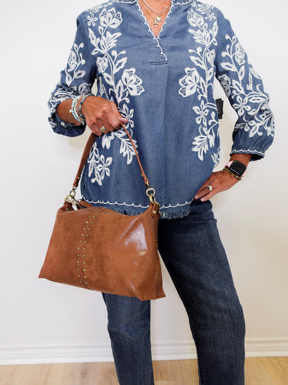 Person wearing a blue floral embroidered shirt and holding a brown handbag against a white background