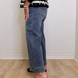 Person wearing a black and white patterned top, blue jeans, and black slippers on a light wooden floor.