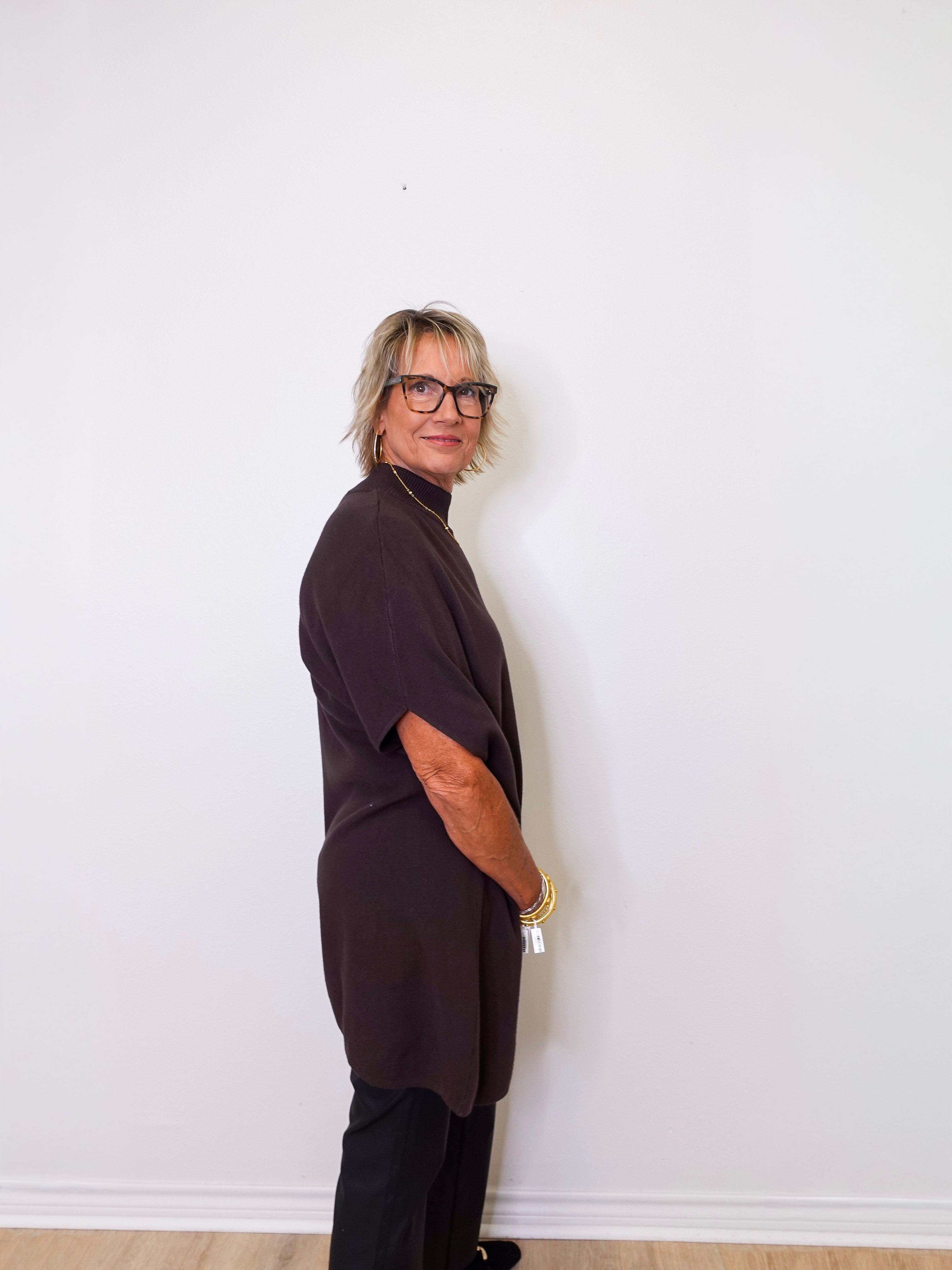 Person wearing a dark brown top and black pants against a white wall.