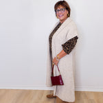 Woman wearing a long beige cardigan over a patterned top, holding a red handbag against a white wall.