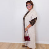 Woman wearing a long beige cardigan over a patterned top, holding a red handbag against a white wall.