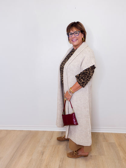 Woman wearing a long beige cardigan over a patterned top, holding a red handbag against a white wall.
