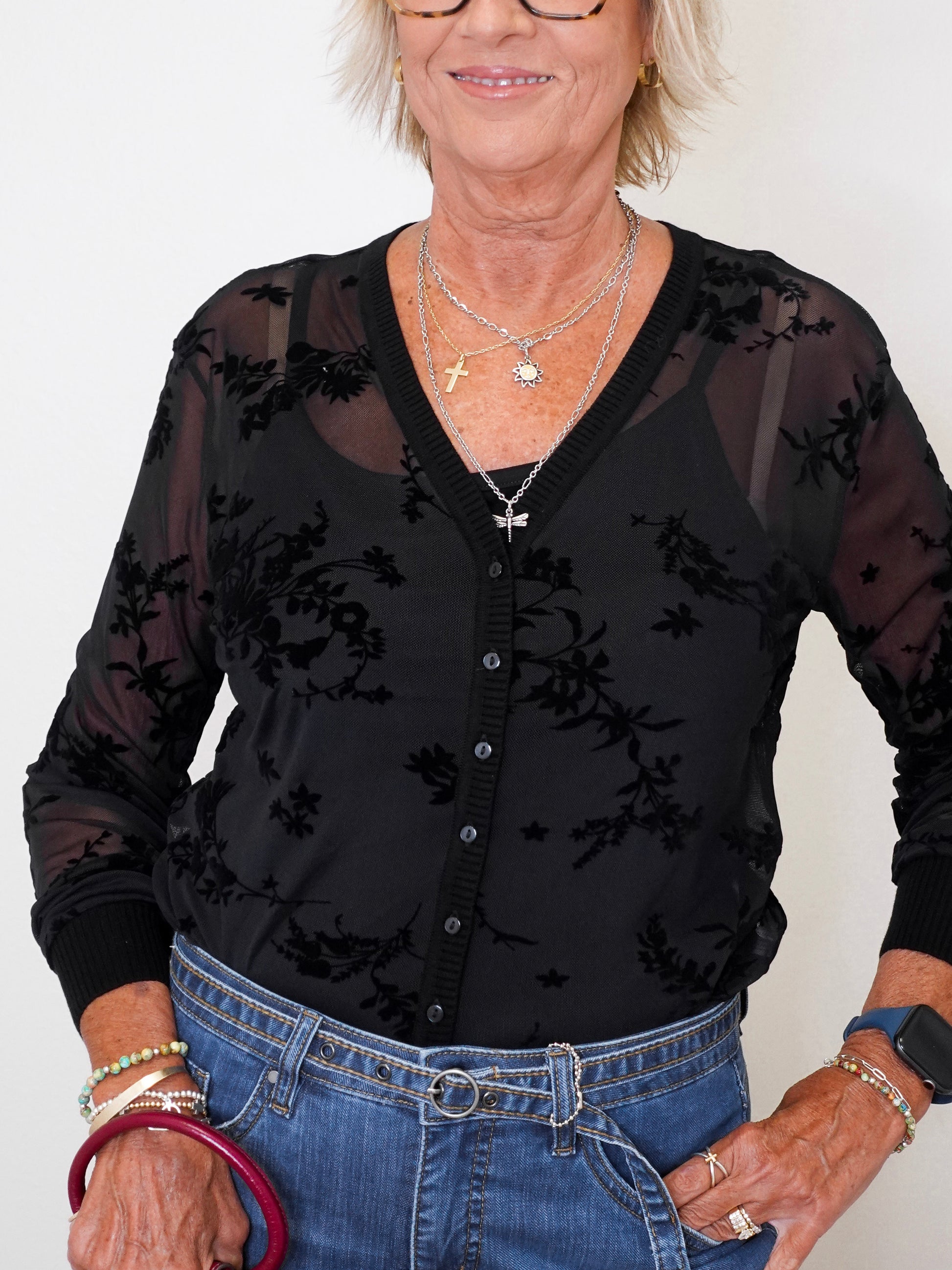 Casual chic outfit: woman in a black mesh cardigan and tank set with blue jeans and a maroon purse, standing against white background.