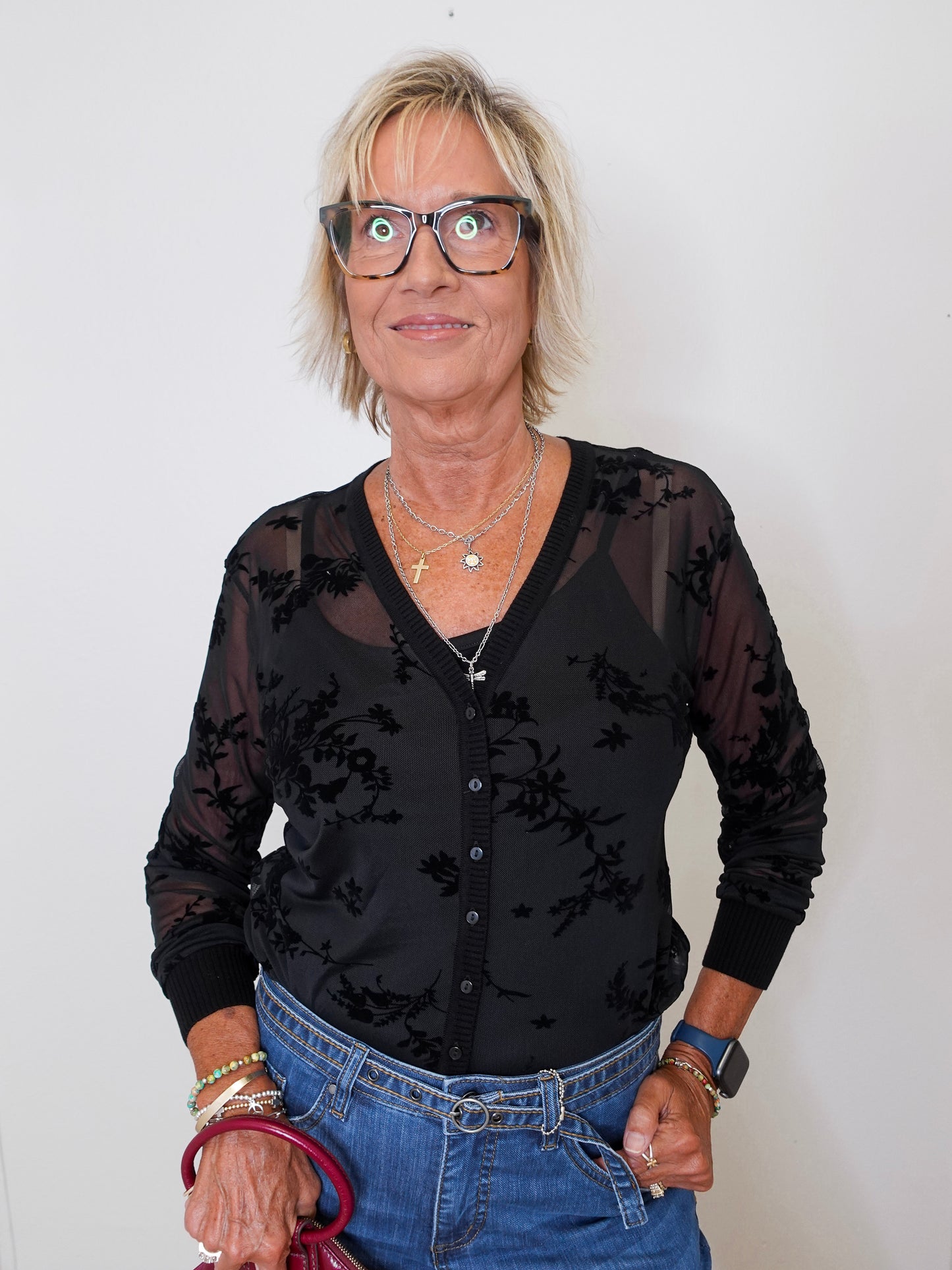 Model wearing a black mesh cardigan and tank set, blue jeans, and holding a maroon purse against a white backdrop.