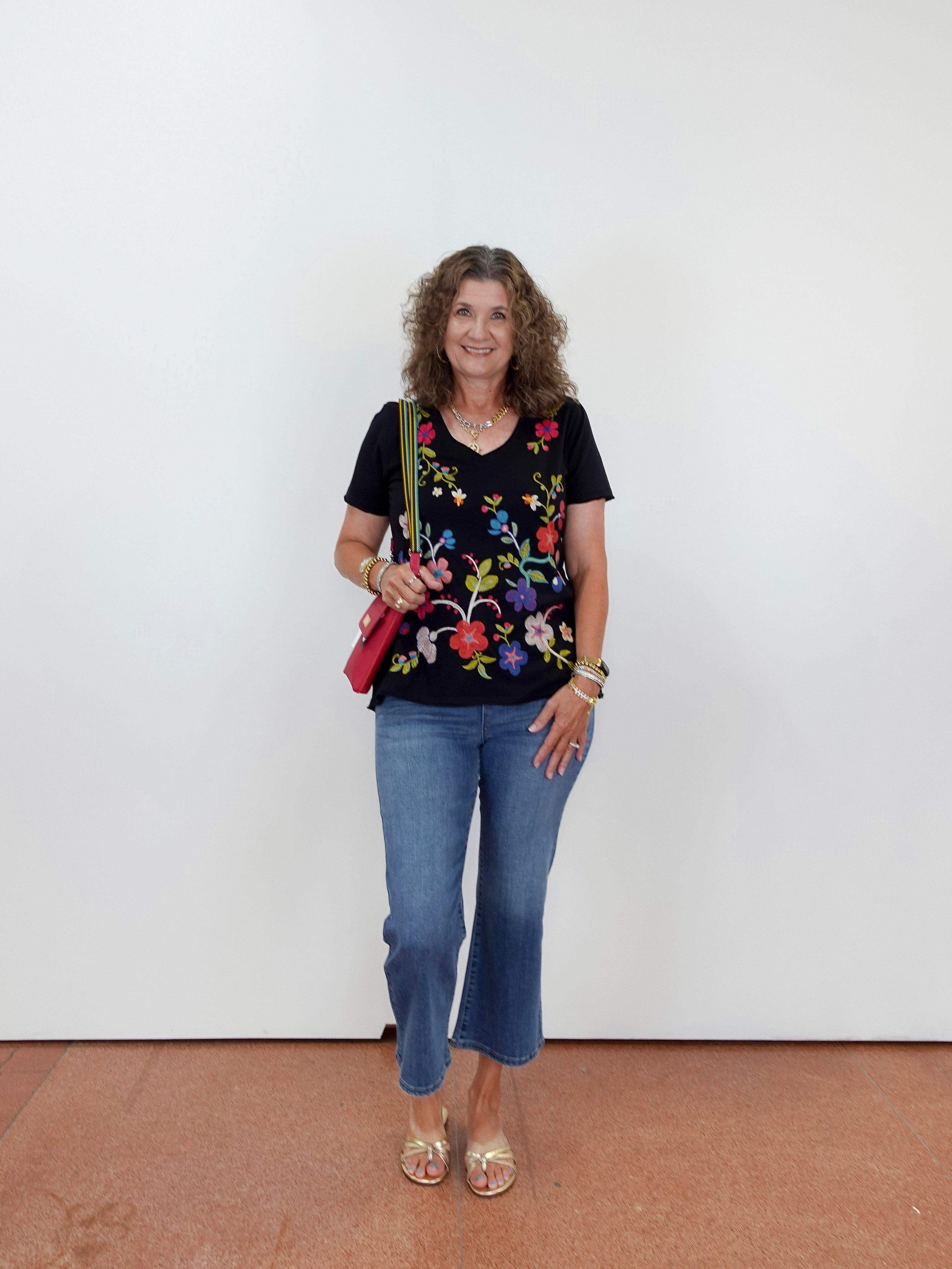 Woman wearing a black floral top and blue jeans against a white wall.