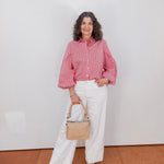 Woman wearing a pink shirt and white pants, holding a beige handbag against a white wall.