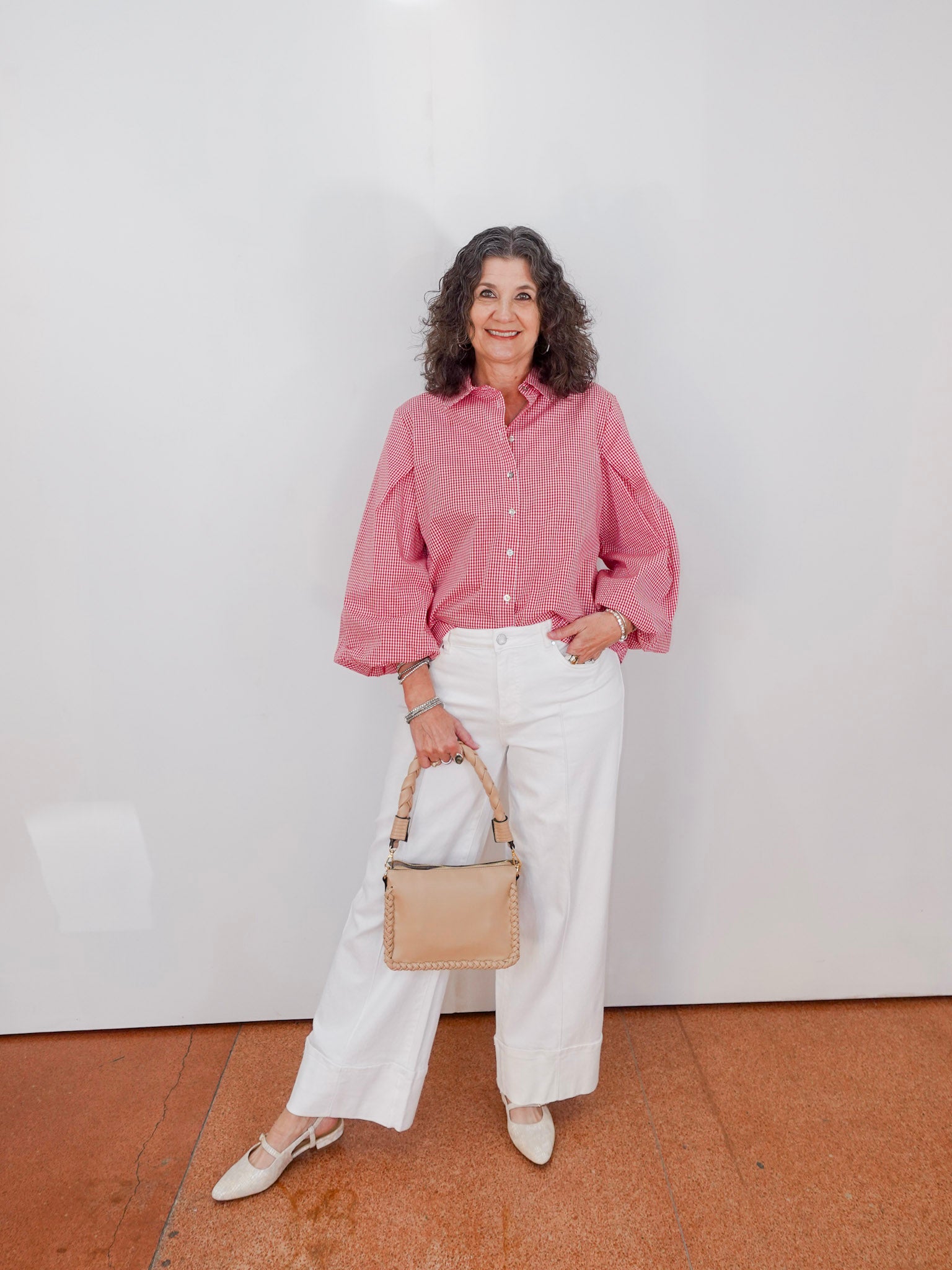 Woman wearing a pink shirt and white pants, holding a beige handbag against a white wall.