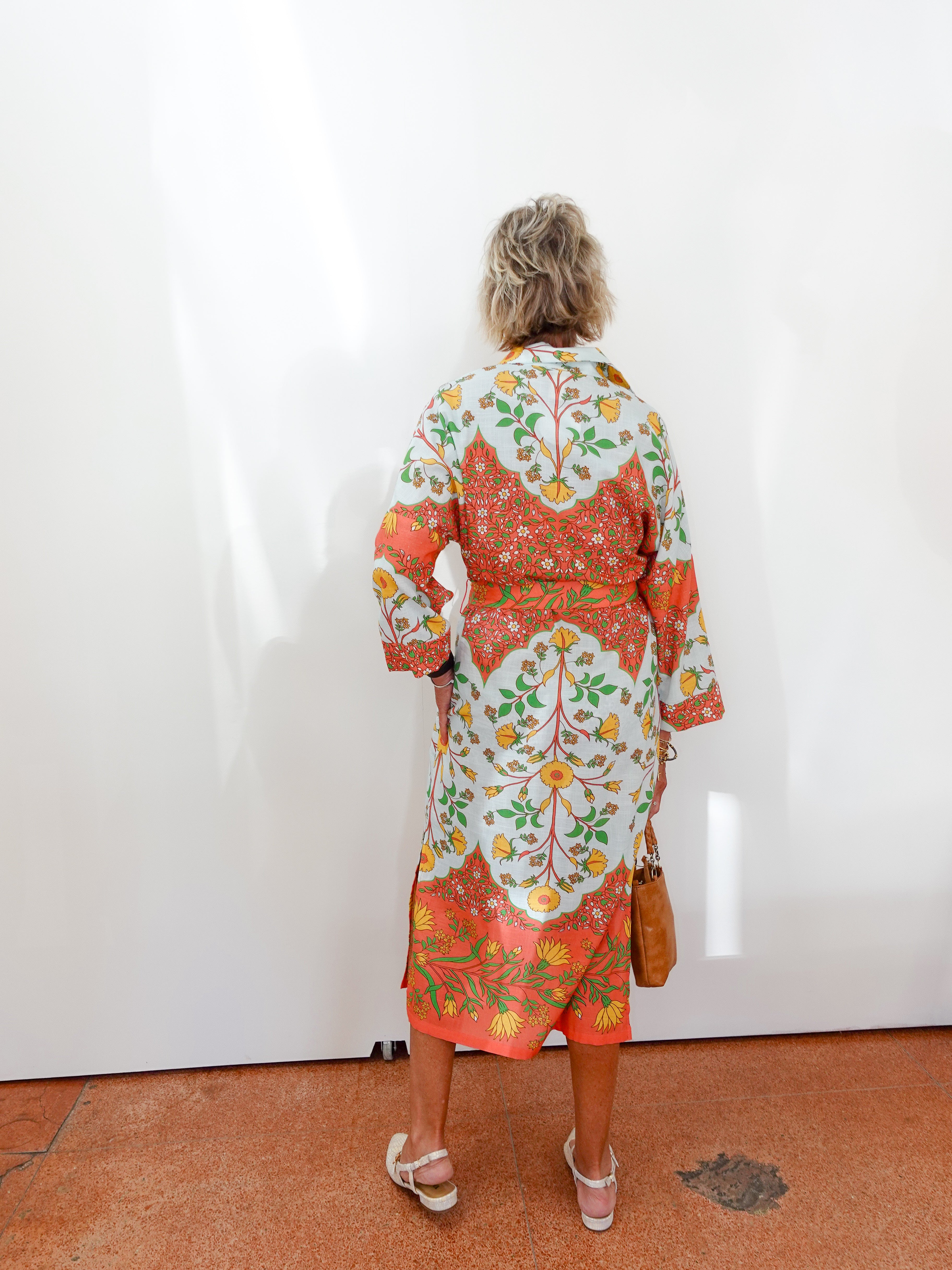 Woman wearing a colorful floral dress standing against a white wall.