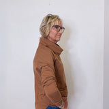 Woman in a tan cowl neck sweater paired with blue jeans standing against a white wall.