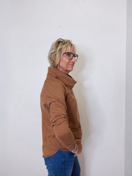 Woman in a tan cowl neck sweater paired with blue jeans standing against a white wall.