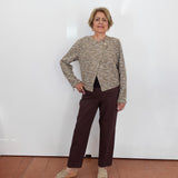 An older woman stands against a white wall wearing a textured beige and brown cropped jacket, dark brown trousers, and woven wedge sandals.