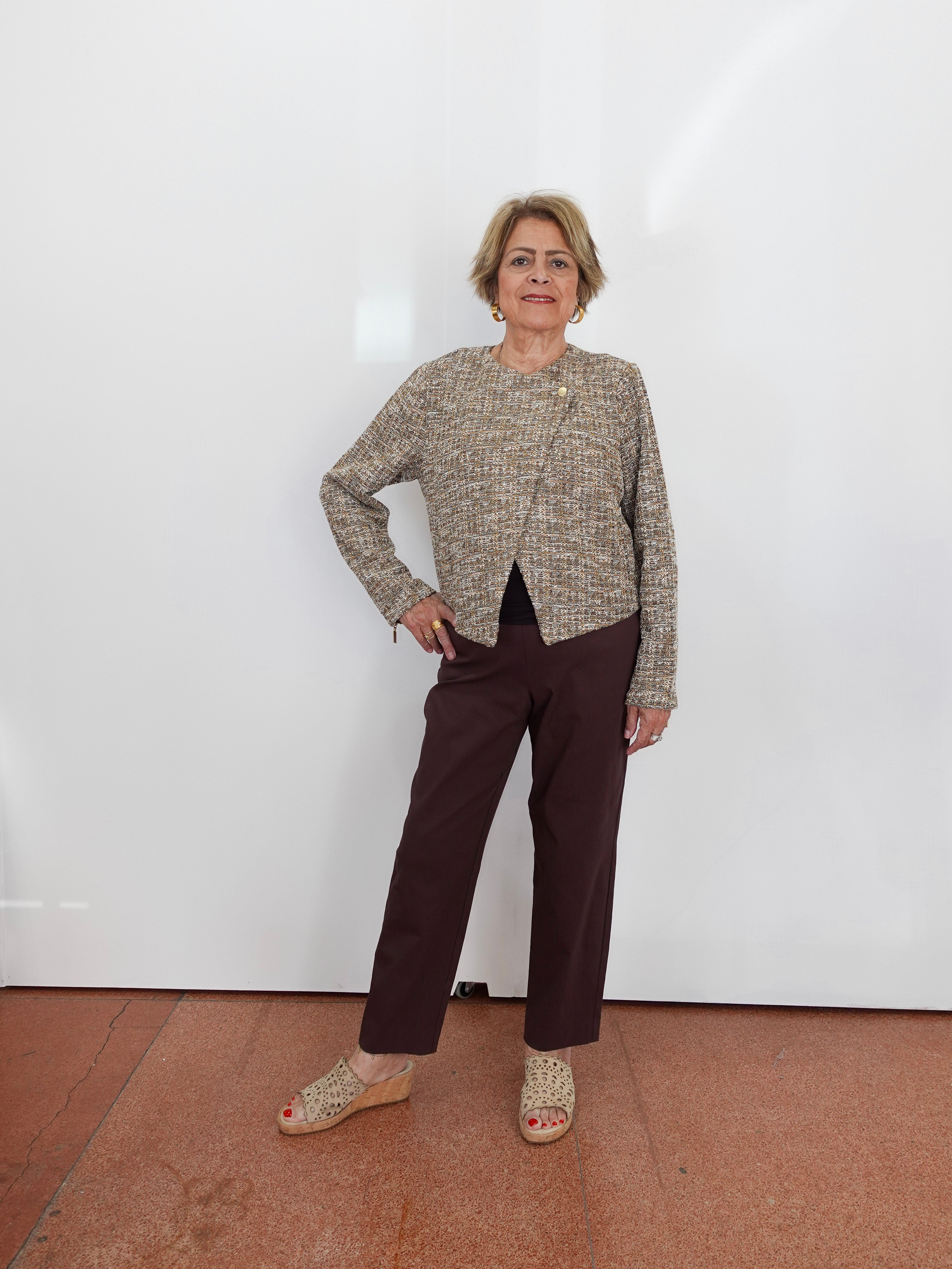 An older woman stands against a white wall wearing a textured beige and brown cropped jacket, dark brown trousers, and woven wedge sandals.