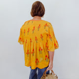 Person wearing a yellow patterned blouse with blue pants against a white wall.