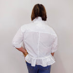 Person wearing a white blouse with ruffled details and blue jeans against a plain background