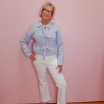 Woman wearing a light blue patterned blouse and white pants against a pink wall.