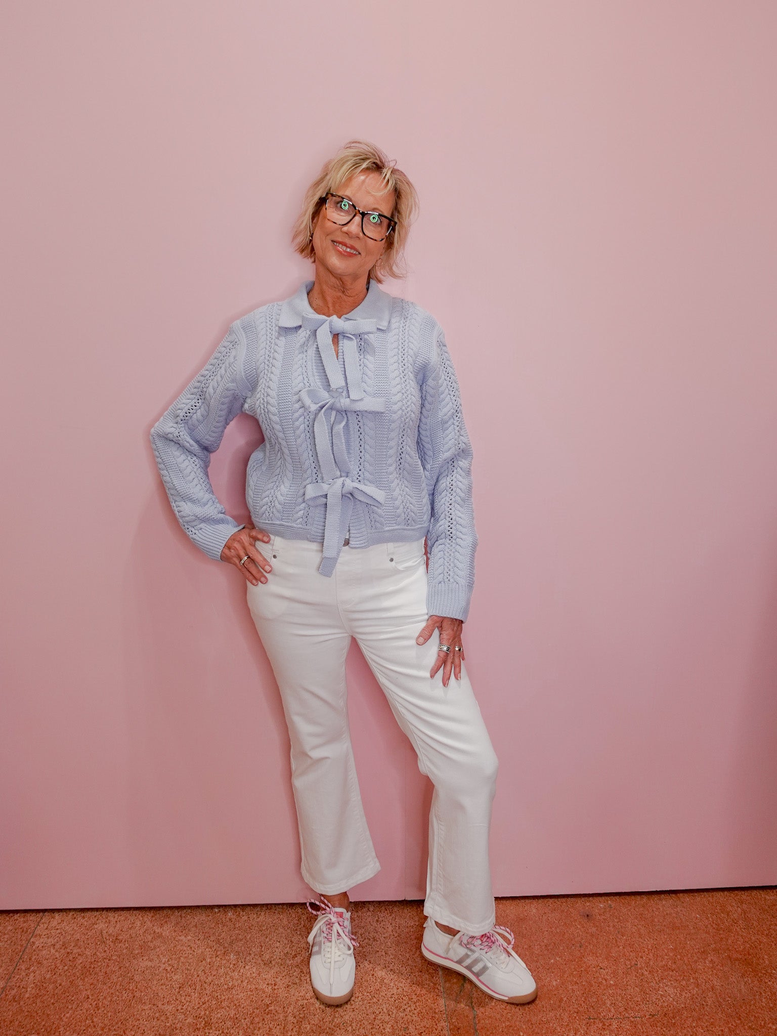 Woman wearing a light blue patterned blouse and white pants against a pink wall.