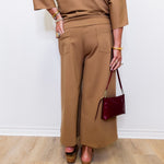 Person wearing a brown outfit with wide-leg pants and holding a red handbag against a white wall.