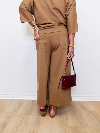Person wearing a brown outfit with wide-leg pants and holding a red handbag against a white wall.