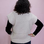 Person wearing a light gray knitted vest over a black long-sleeve shirt against a pink background