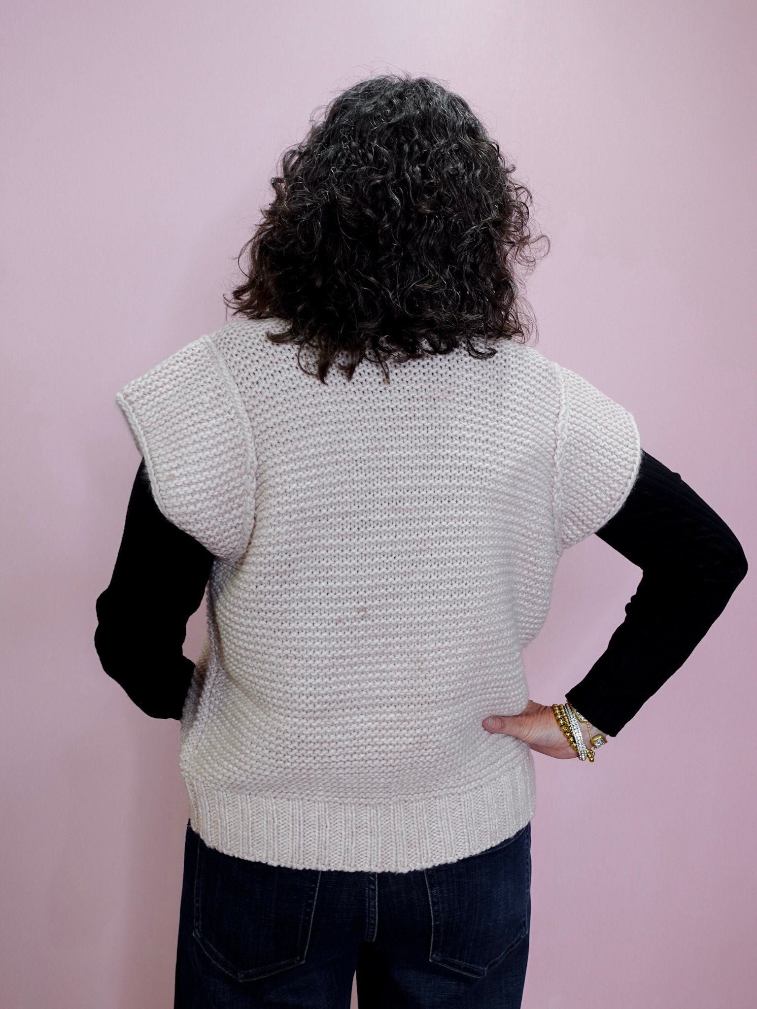 Person wearing a light gray knitted vest over a black long-sleeve shirt against a pink background