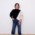 Woman wearing a black and beige sweater with blue jeans against a white wall.
