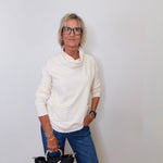 A woman wearing a cowl neck sweater in cream with blue pants against a white wall. 