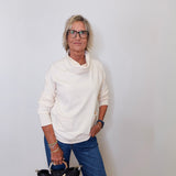 A woman wearing a cowl neck sweater in cream with blue pants against a white wall. 