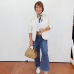 Woman in a white blazer, blue jeans, and white sneakers holding a beige handbag against a white wall.
