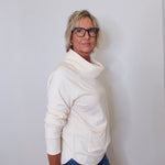 A woman wearing a cowl neck sweater in cream with blue pants against a white wall. 
