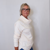 A woman wearing a cowl neck sweater in cream with blue pants against a white wall. 