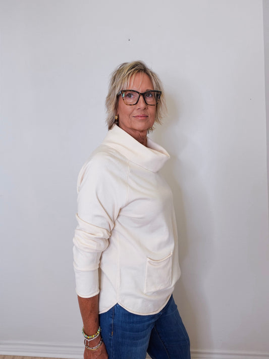 A woman wearing a cowl neck sweater in cream with blue pants against a white wall. 