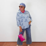 Woman wearing a blue patterned jacket and jeans, holding a pink handbag against a white wall.