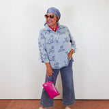 Woman wearing a blue patterned jacket and jeans, holding a pink handbag against a white wall.