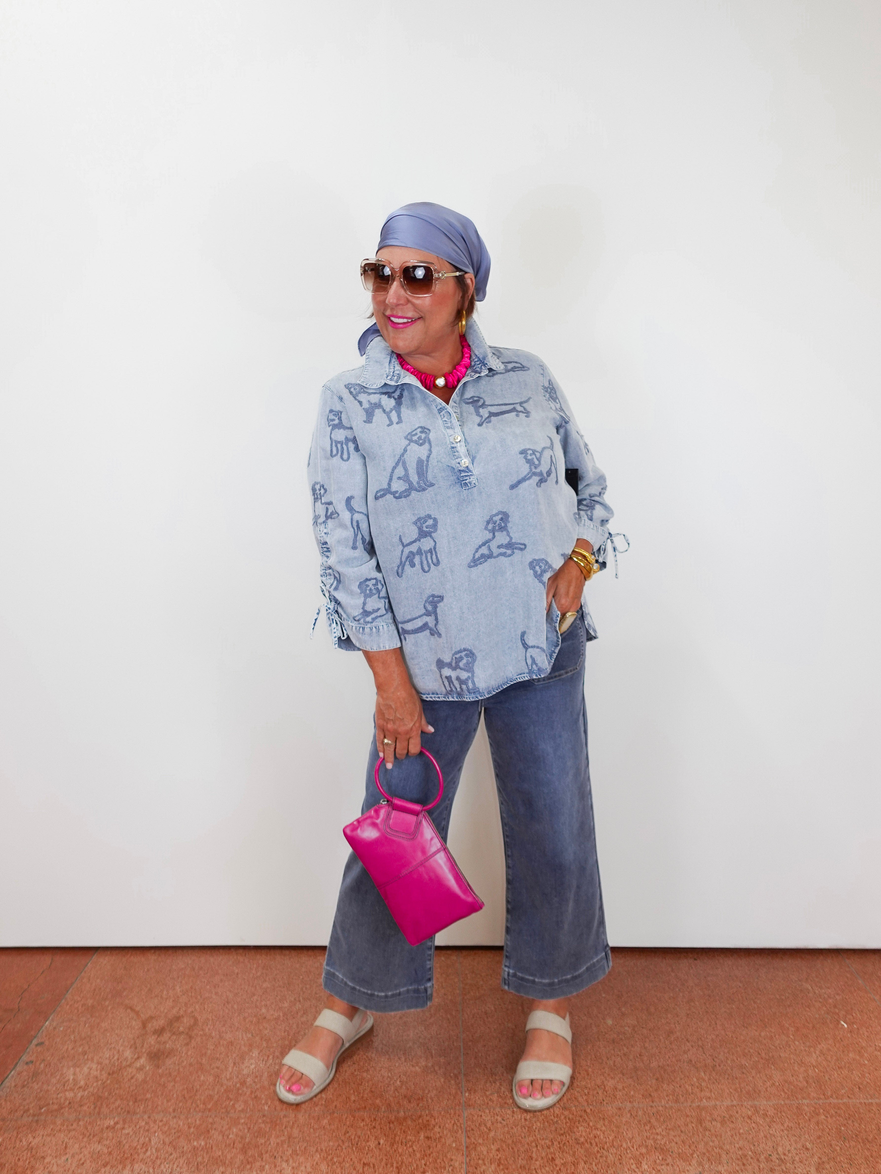 Woman wearing a blue patterned jacket and jeans, holding a pink handbag against a white wall.