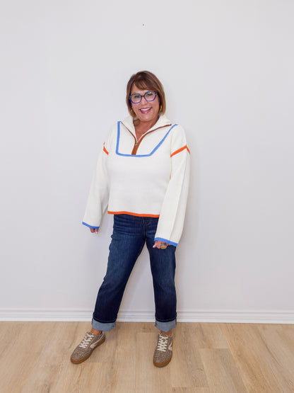 Person wearing a colorful sweater and jeans standing against a white wall.
