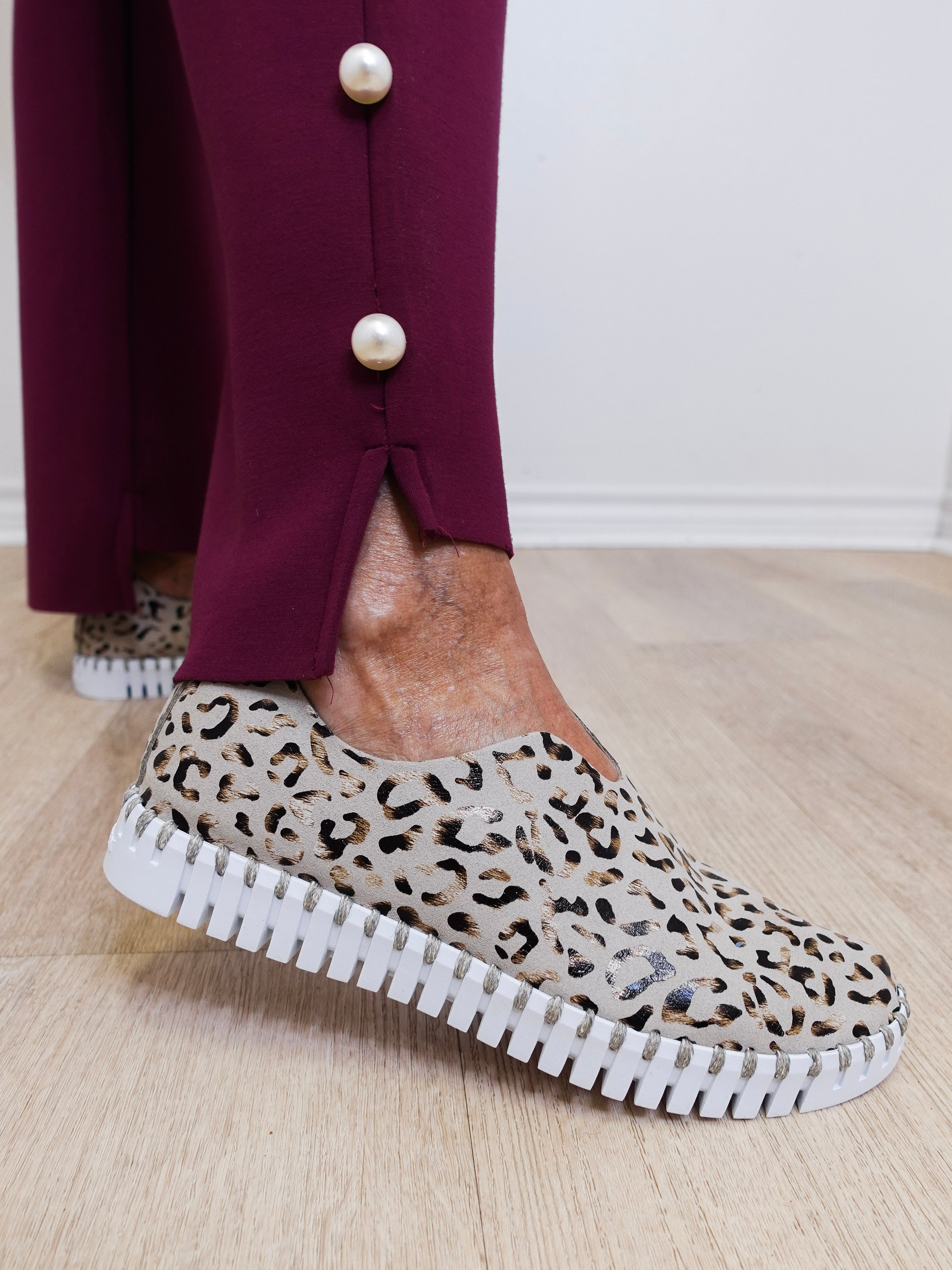 Person wearing leopard print slip-on shoes with a white sole on a wooden floor.
