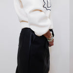 Person wearing a white sweater and black skirt against a white wall