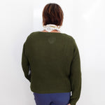A woman viewed from behind wearing an olive green textured knit sweater, blue pants, and a cream scarf, holding a brown leather bag against a plain white background.