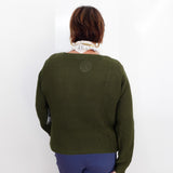 A woman viewed from behind wearing an olive green textured knit sweater, blue pants, and a cream scarf, holding a brown leather bag against a plain white background.