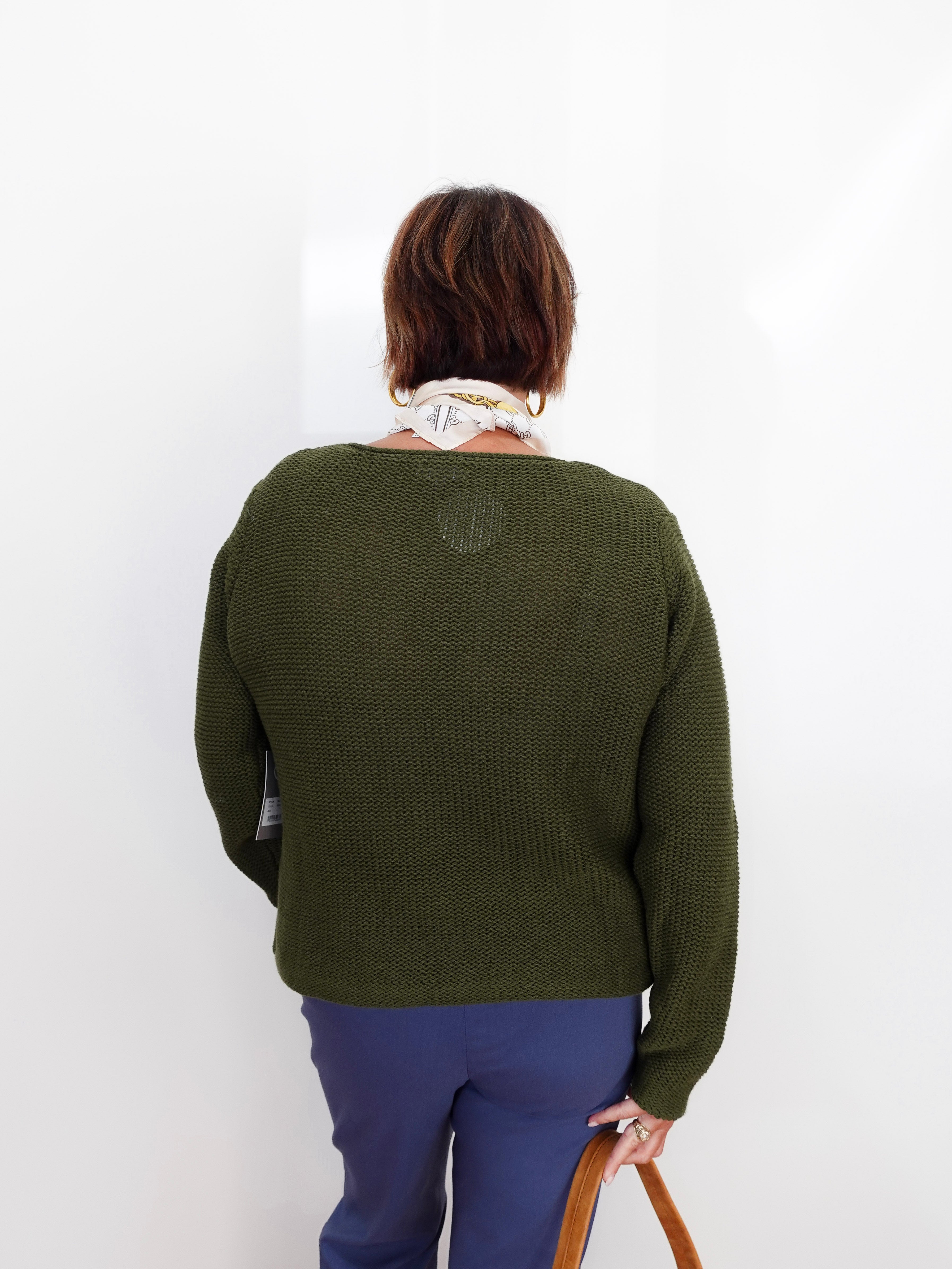 A woman viewed from behind wearing an olive green textured knit sweater, blue pants, and a cream scarf, holding a brown leather bag against a plain white background.