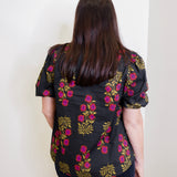 Person wearing a black floral blouse with pink and yellow flowers against a white wall.
