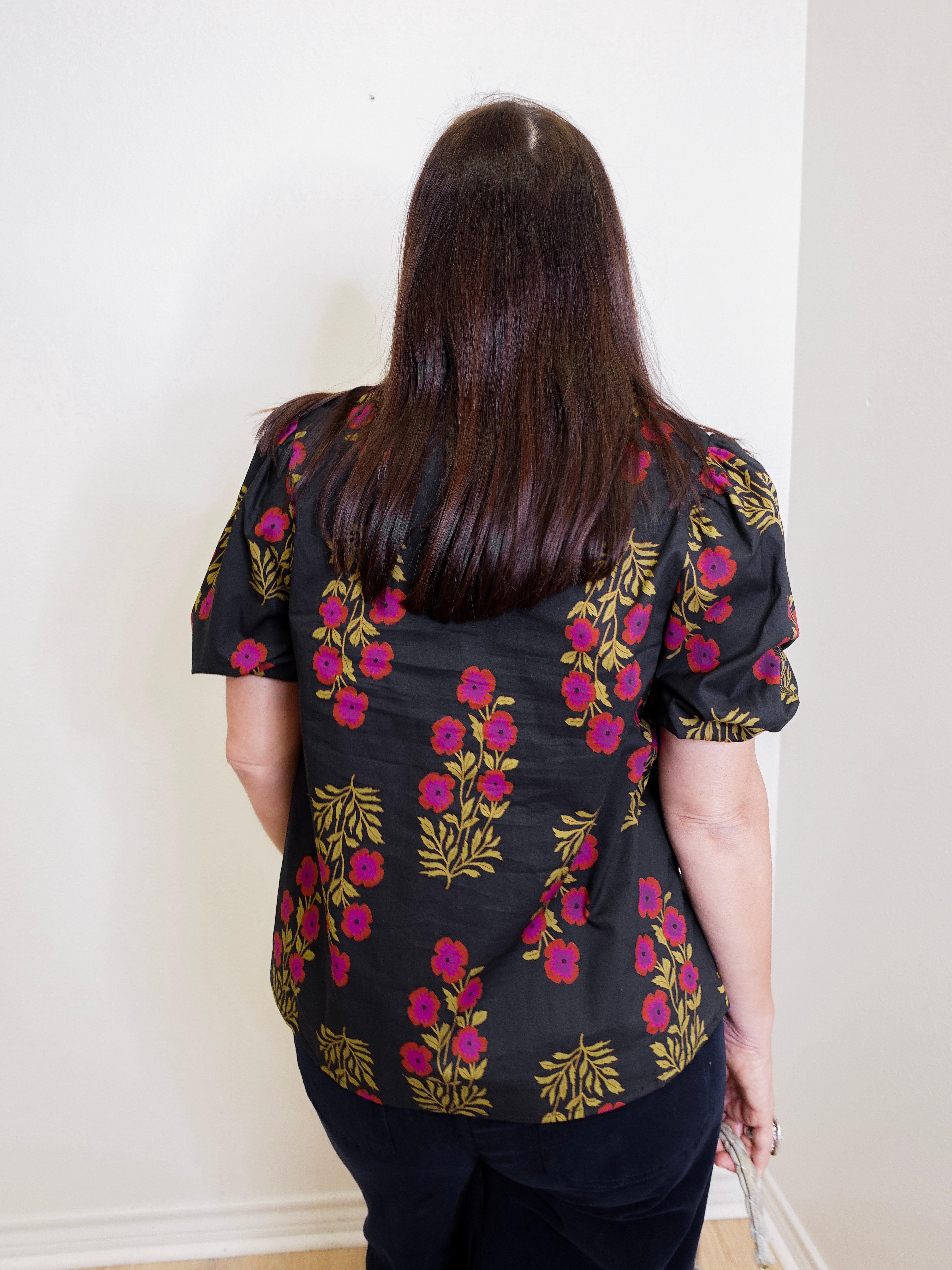 Person wearing a black floral blouse with pink and yellow flowers against a white wall.