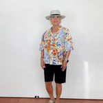 Person wearing a colorful floral shirt, black shorts, and a hat against a white wall.