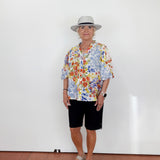 Person wearing a colorful floral shirt, black shorts, and a hat against a white wall.