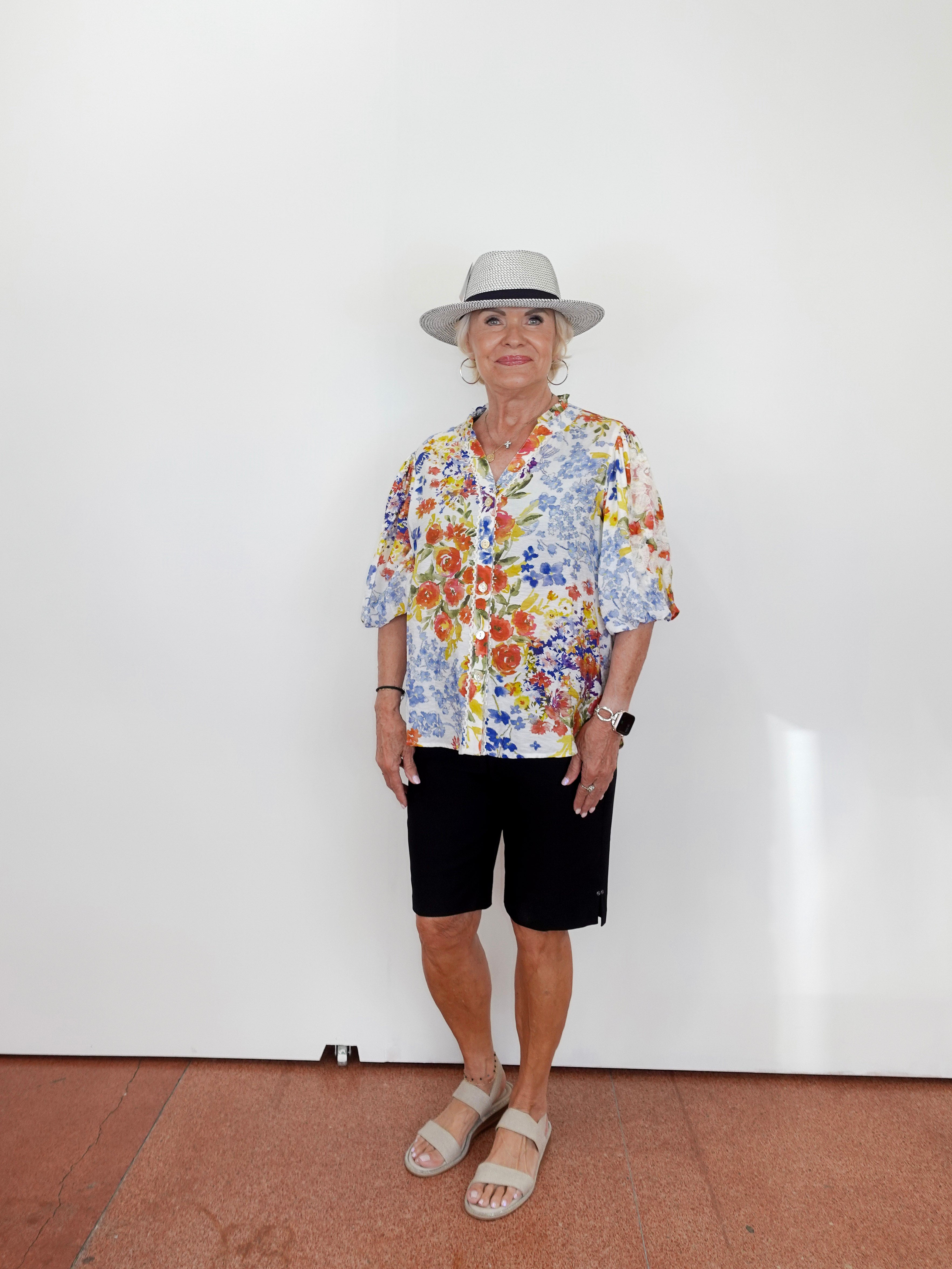 Person wearing a colorful floral shirt, black shorts, and a hat against a white wall.