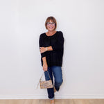 Woman wearing a black sweater and blue jeans, holding a patterned handbag against a white wall.