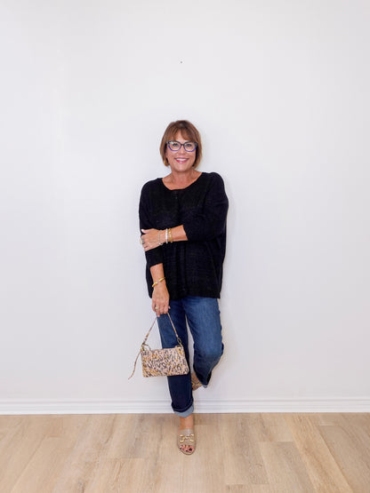 Woman wearing a black sweater and blue jeans, holding a patterned handbag against a white wall.