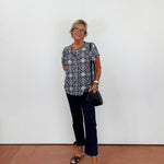 Woman wearing a patterned shirt and dark pants against a white wall.