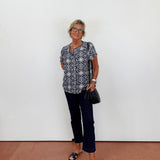 Woman wearing a patterned shirt and dark pants against a white wall.