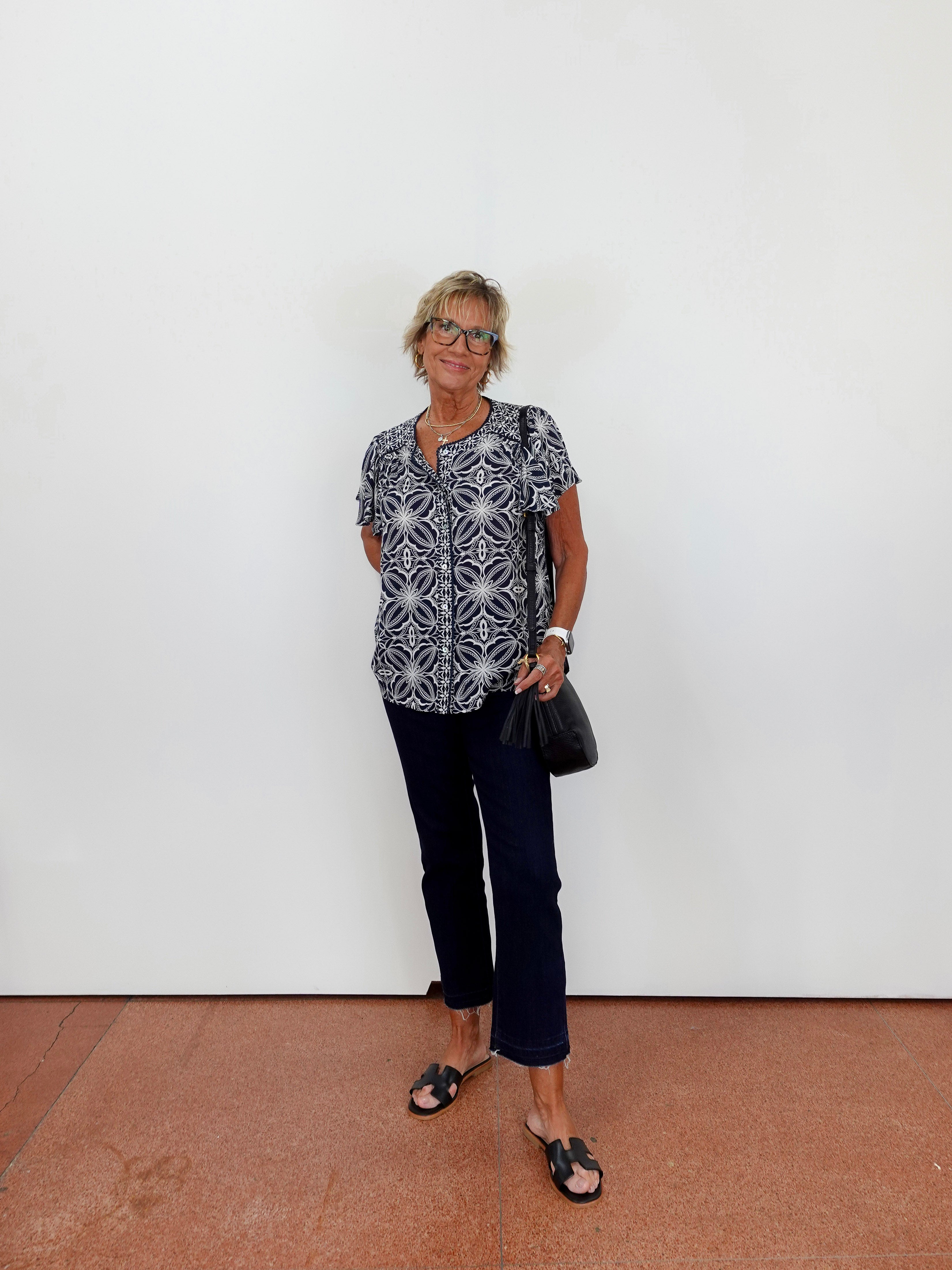 Woman wearing a patterned shirt and dark pants against a white wall.